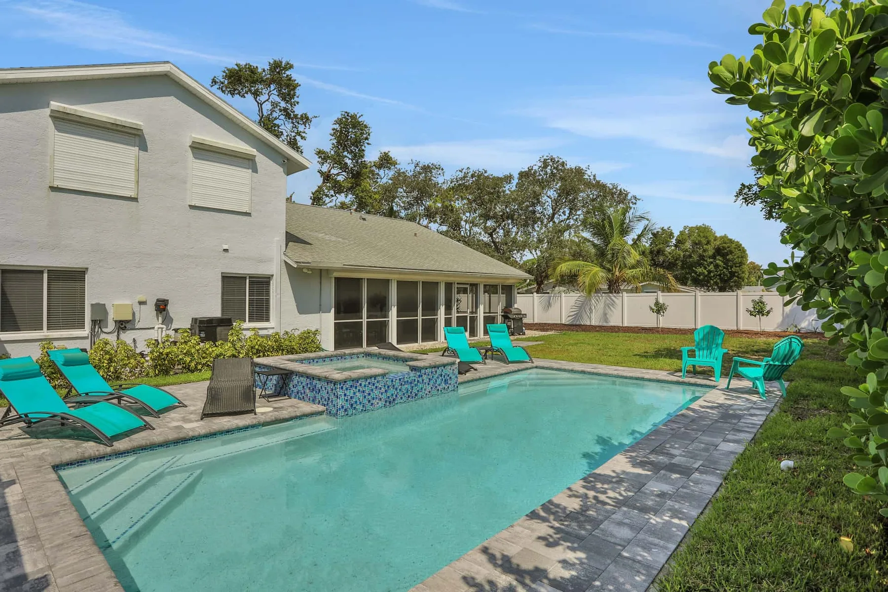 A backyard swimming pool sits beside a house, with lounge chairs and a fenced lawn.