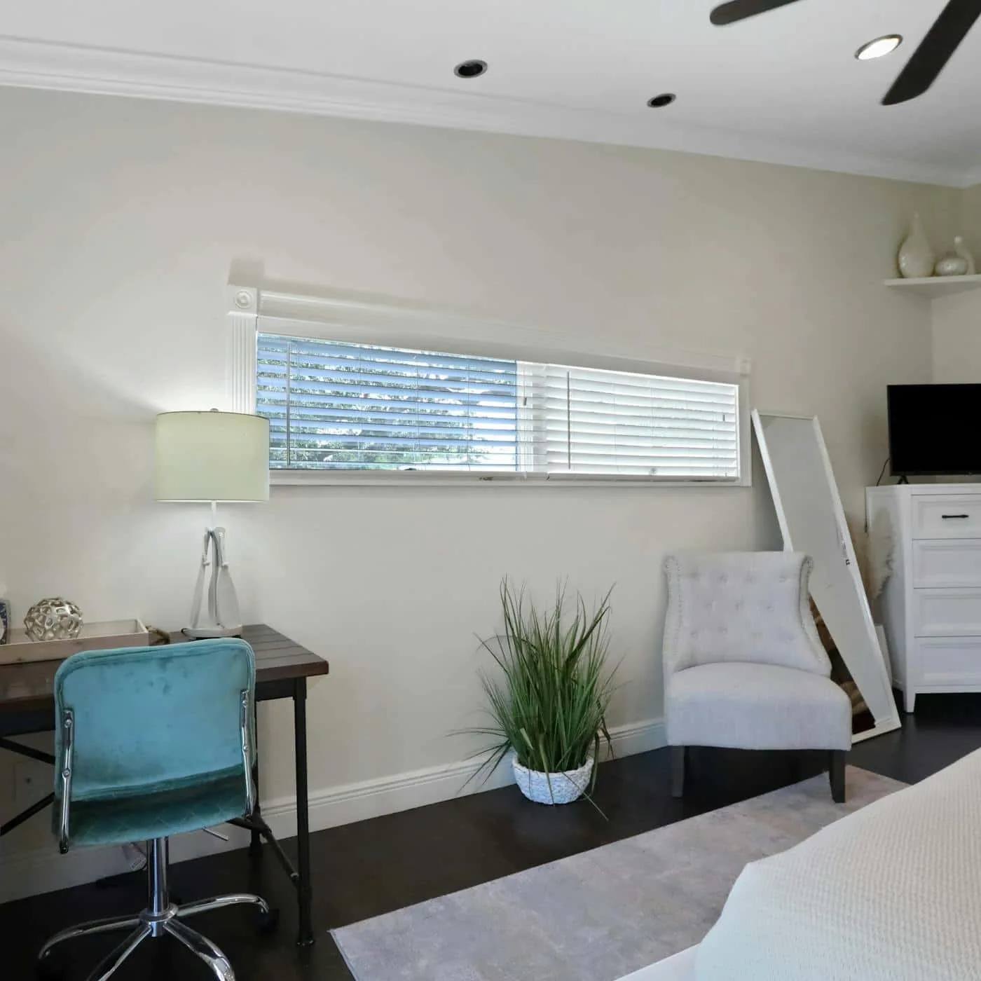 A bright bedroom with a desk, a white chair, and a long window with blinds.