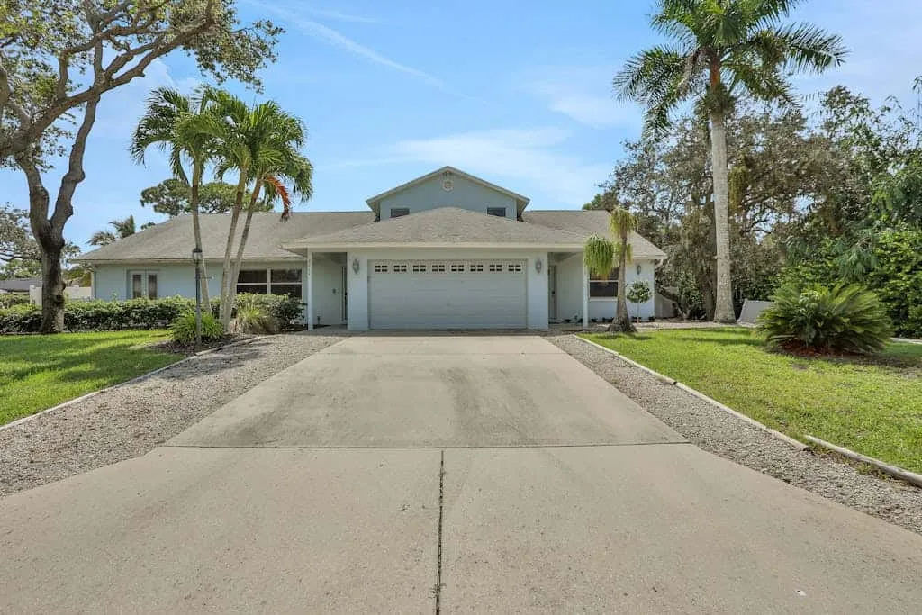 A single-story house faces a wide concrete driveway, with palm trees on the lawn.
