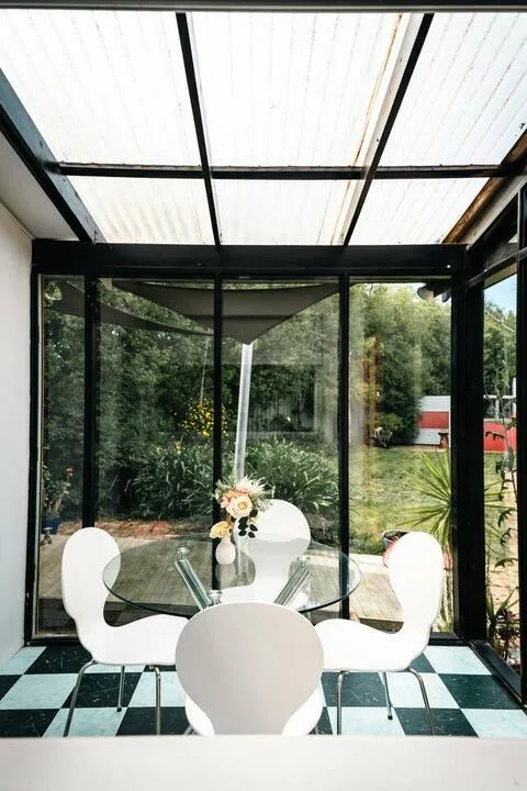 A glass-enclosed dining area with a round table and white seating overlooks a garden.