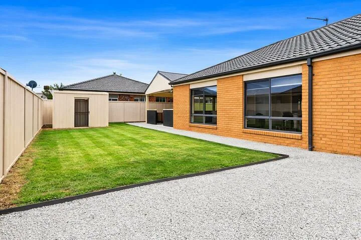 A grassy backyard sits beside a brick house and paved area.