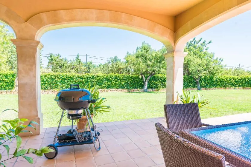 Un patio cubierto da a un jardín verde, con una parrilla y asientos al aire libre.