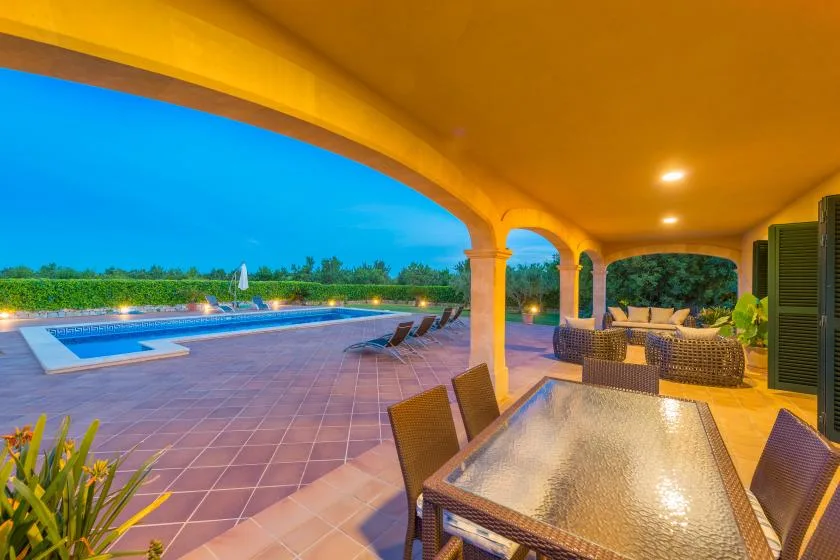 Un patio cubierto con comedor da a una piscina al anochecer.