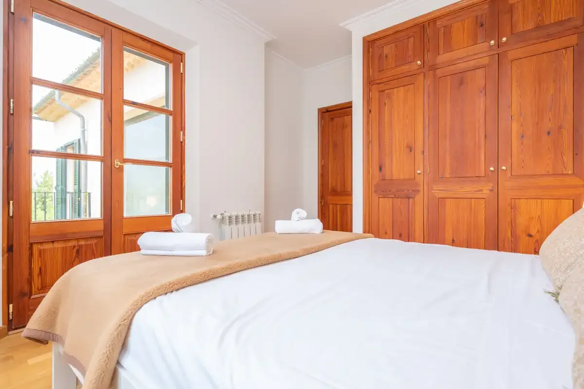 Un dormitorio con una cama bien arreglada junto a armarios de madera altos y ventanas.