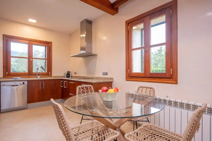 Una luminosa cocina con zona de comedor, muebles de madera, mesa de vidrio y ventanas grandes.