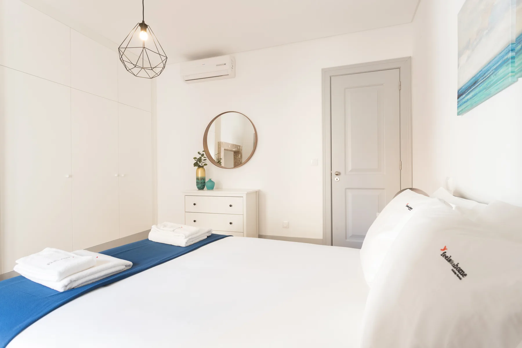 Un dormitorio luminoso con una cama bien arreglada y una cómoda bajo un espejo redondo.