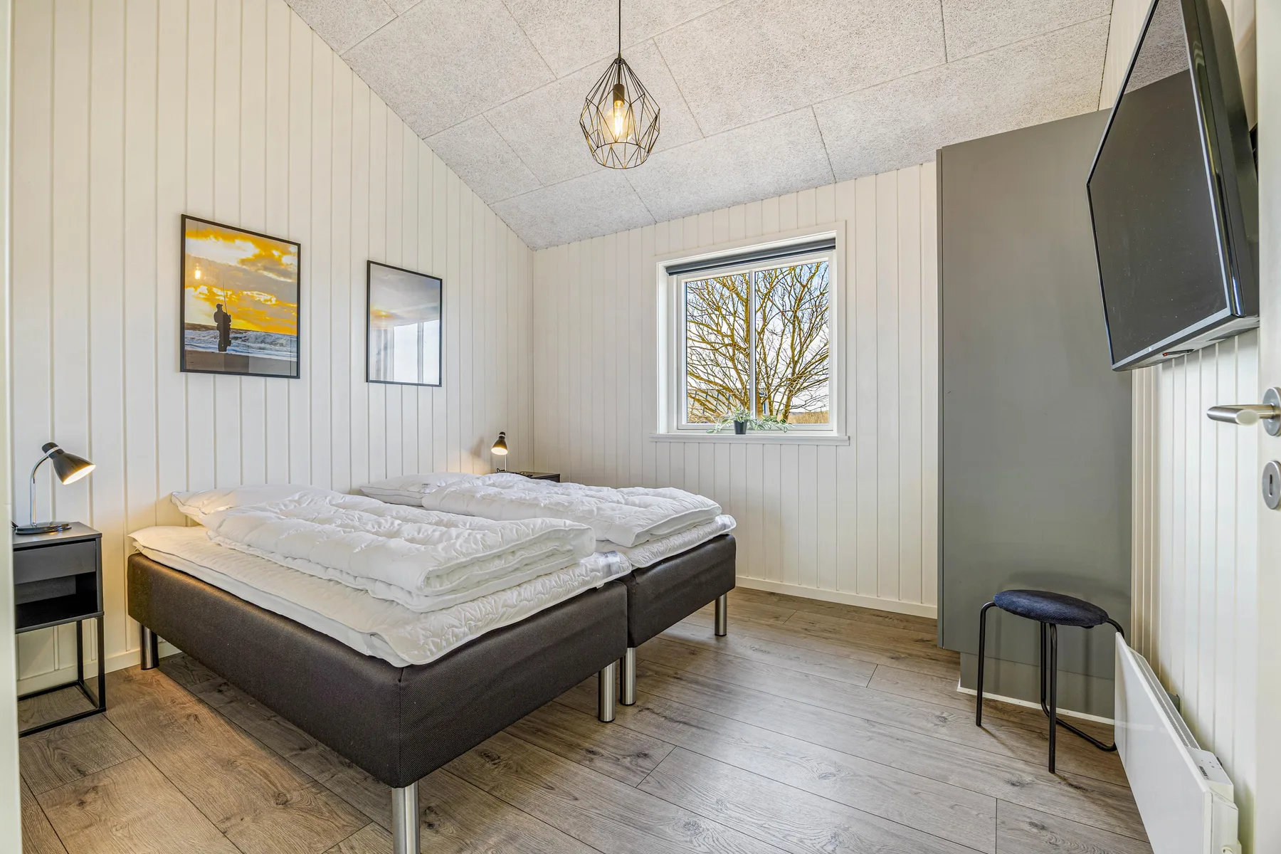 Un dormitorio luminoso con una cama, una ventana y un televisor montado en la pared.