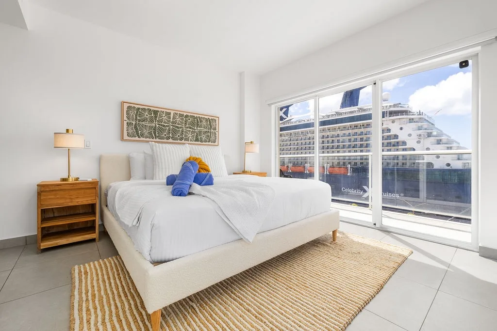 Unwind in your bright bedroom with crisp linens and cruise ship views, where coastal light and warm textures create your restful retreat after exploring.