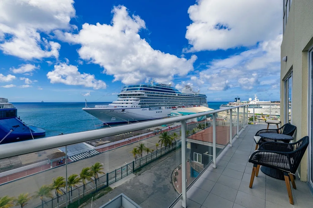 The balcony showcases a panoramic harbor vista with cruise ships and turquoise waters, framed by vibrant blue skies and modern seating for lounging outdoors.