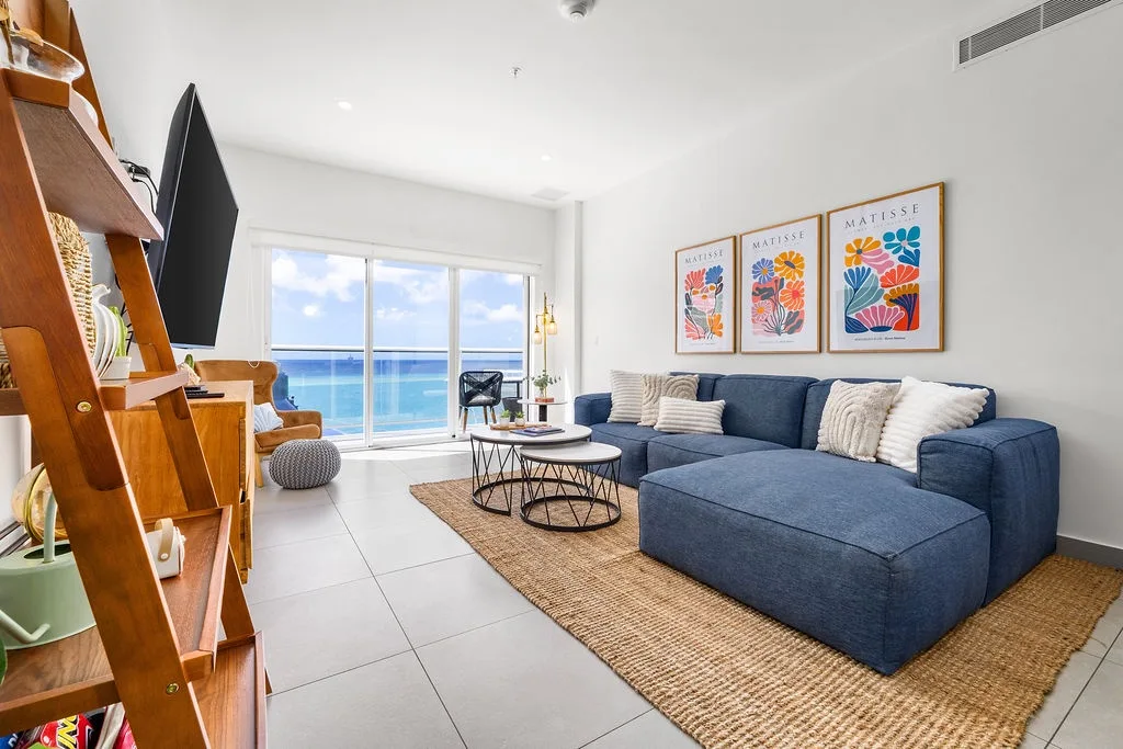 Unwind in your bright, modern living space with ocean views through floor-to-ceiling windows. The plush blue sectional invites relaxation while artwork adds colorful charm.