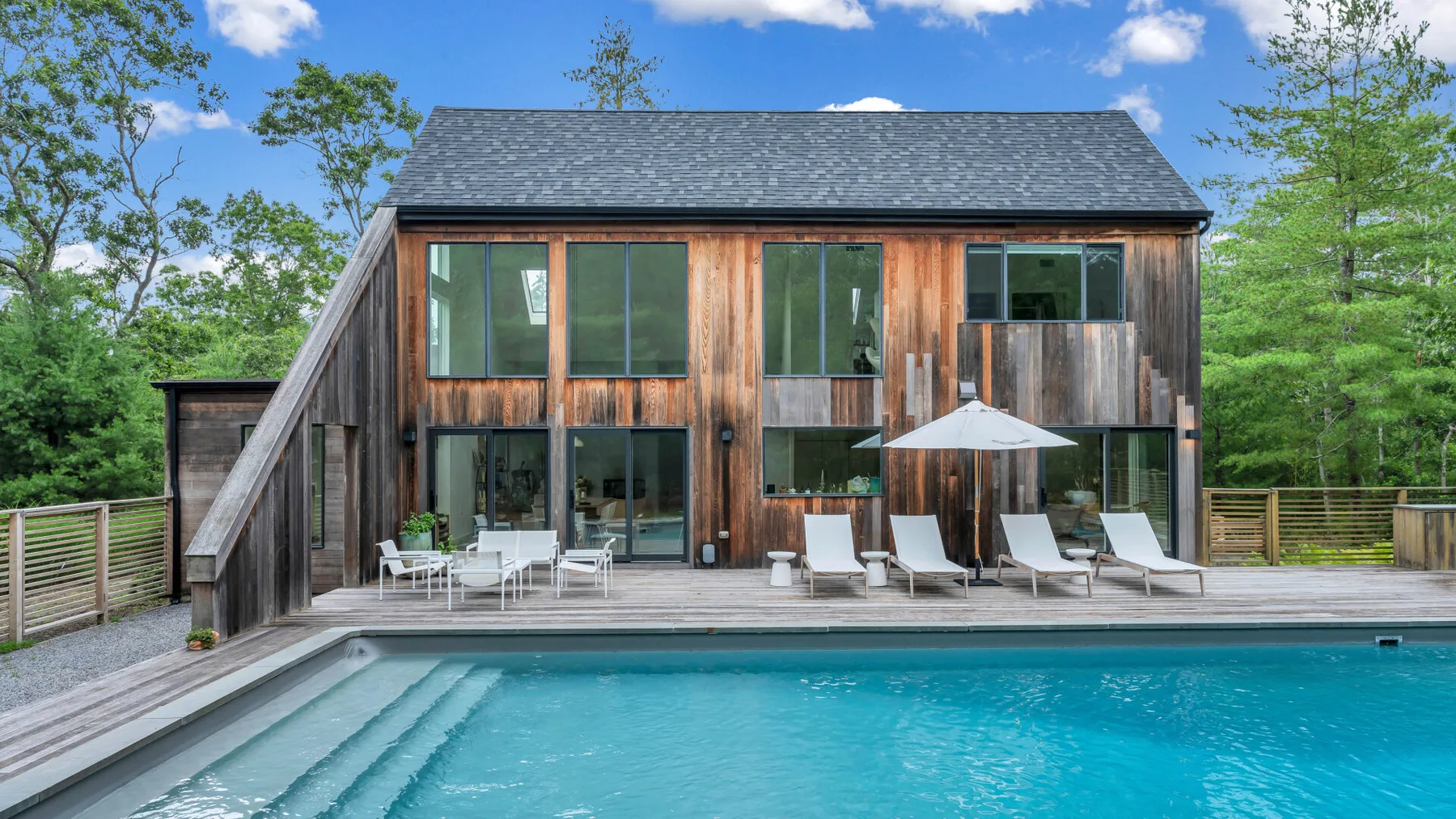 Una casa moderna revestida de madera se alza detrás de una piscina y asientos en el patio.