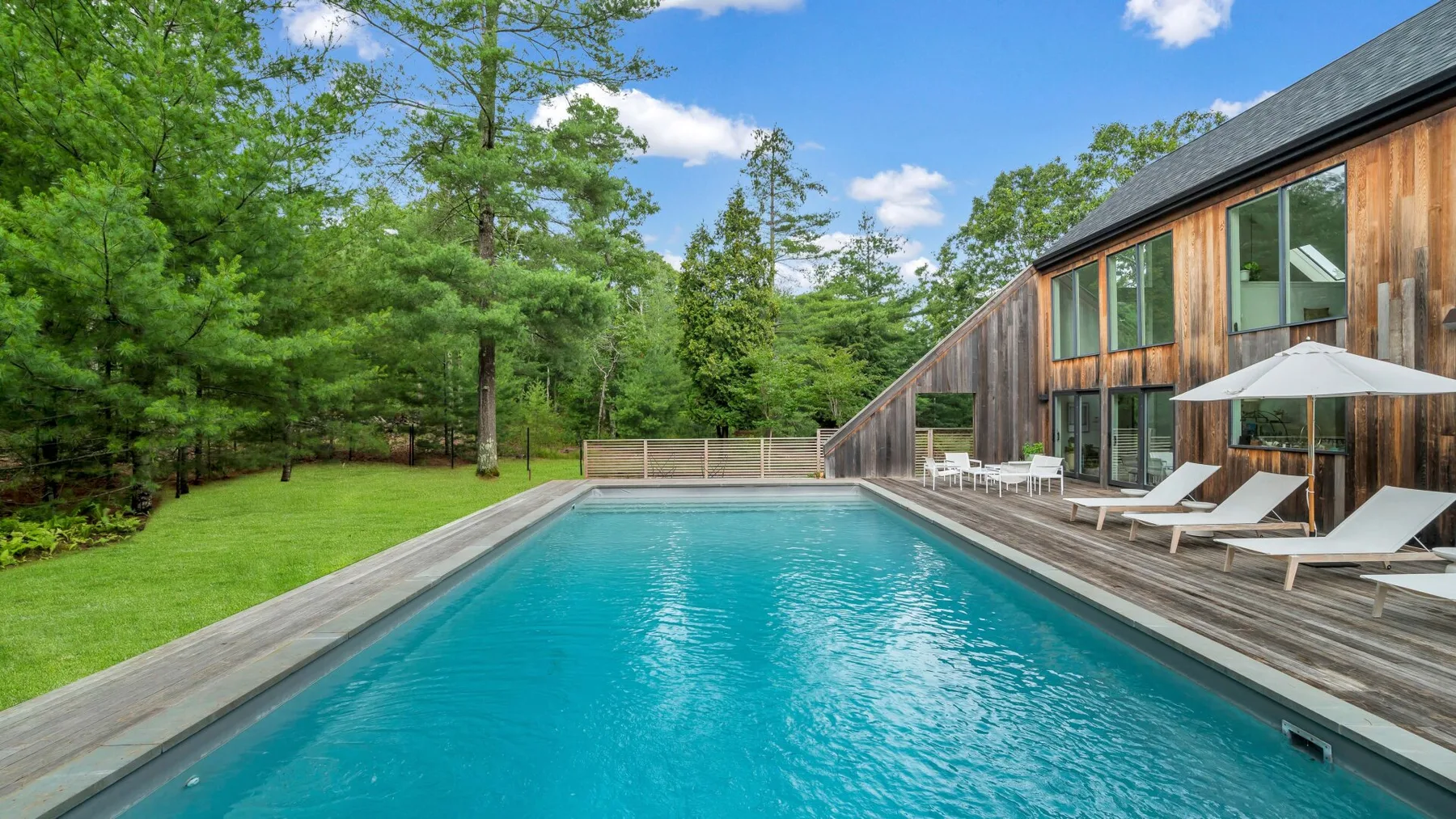 Una piscina exterior junto a una casa de madera, con tumbonas y árboles cerca.