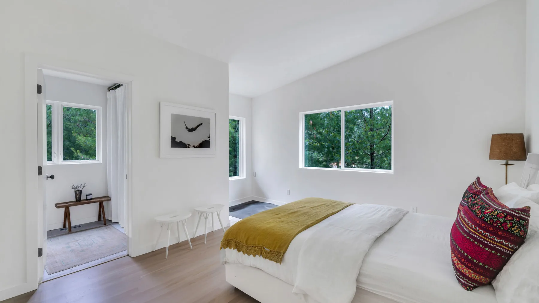 Un dormitorio luminoso con ropa de cama blanca y una manta mostaza junto a grandes ventanas.
