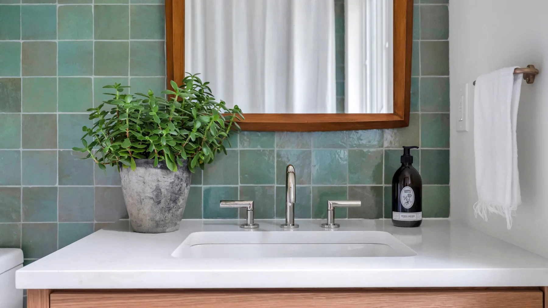 Lavabo de baño con lavabo blanco, pared de azulejos verdes y una planta en maceta.