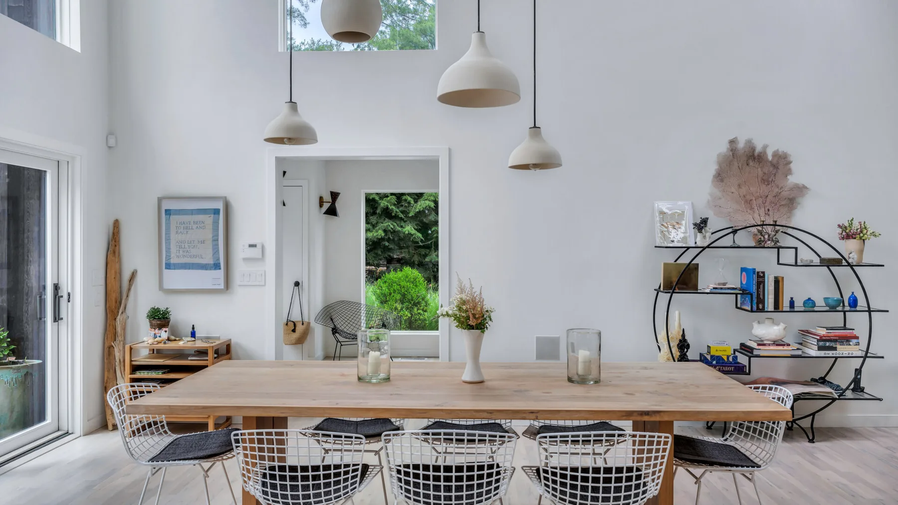 Un comedor luminoso con una mesa larga de madera, asientos de alambre y lámparas colgantes.