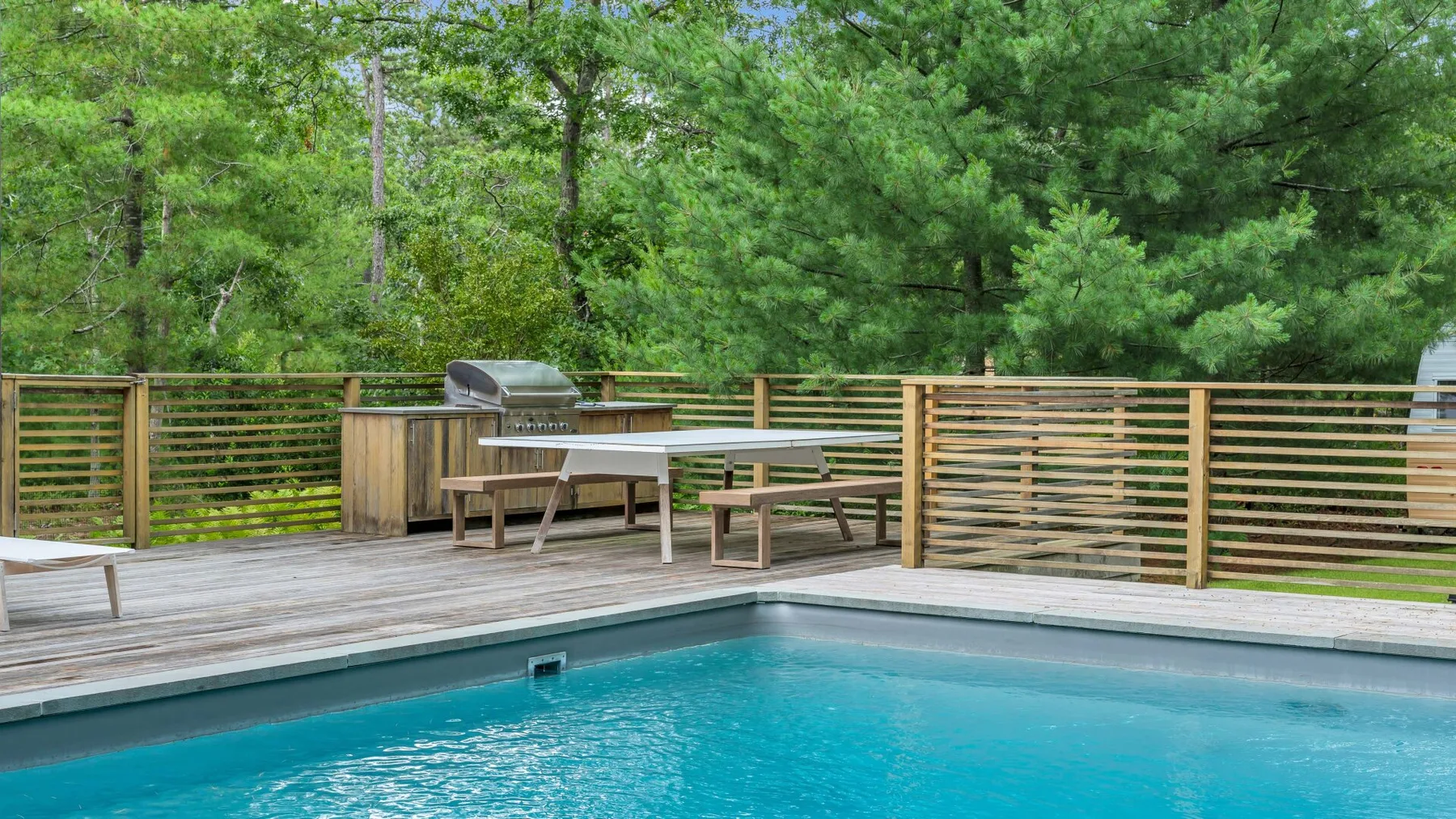 Una piscina al aire libre está junto a una terraza y una barandilla de madera, rodeada de árboles.