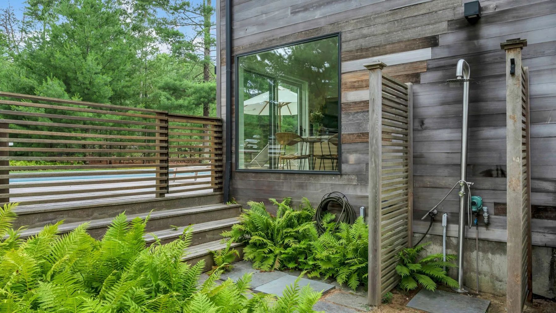 Una ducha exterior junto a una terraza de madera, con helechos y una ventana grande.