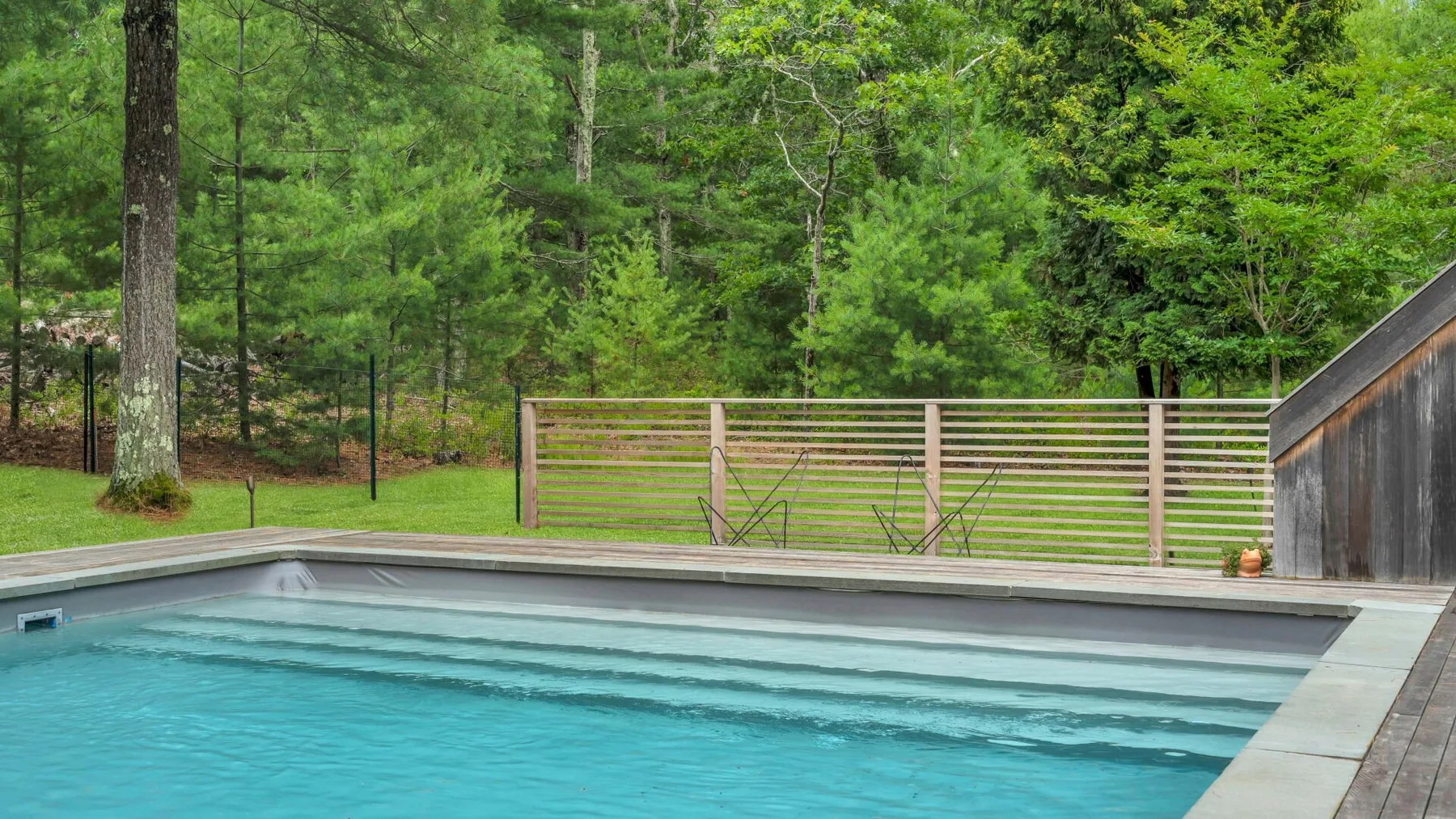 Una piscina al aire libre junto a una terraza de madera, con una zona arbolada al fondo.