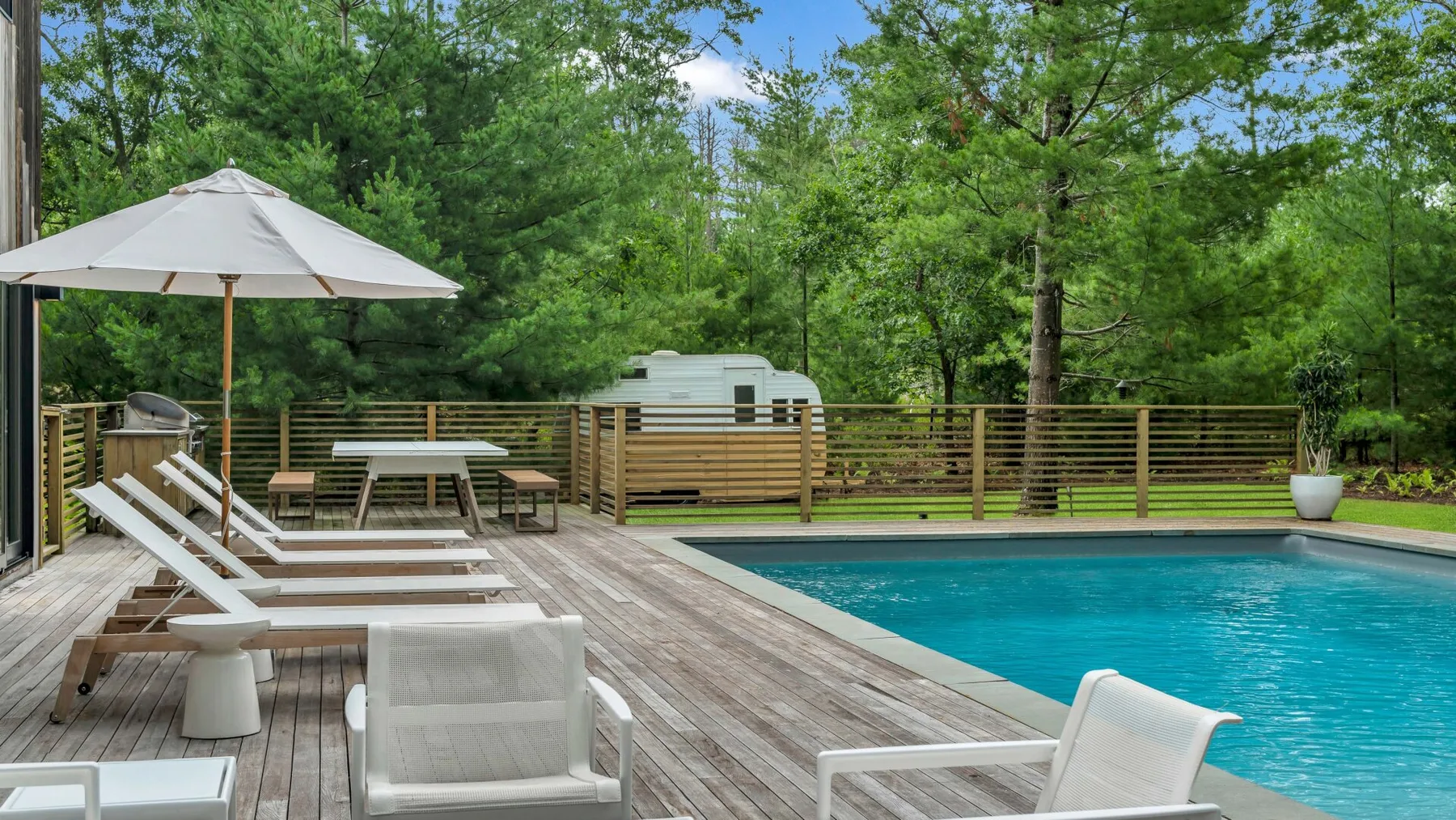 Una piscina al aire libre junto a una terraza de madera, asientos y árboles.