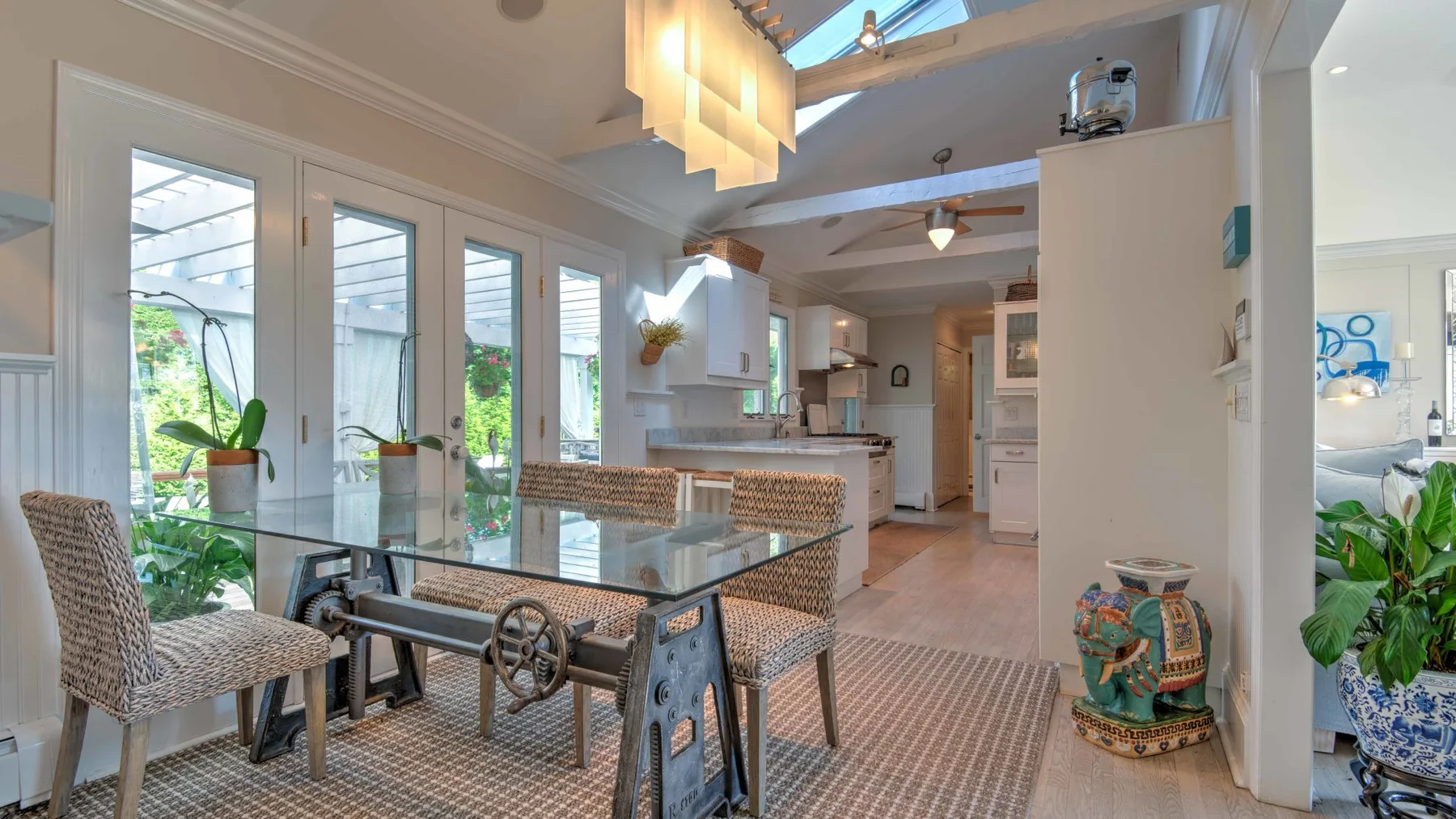 A bright dining area with a glass table beside tall windows and indoor plants.