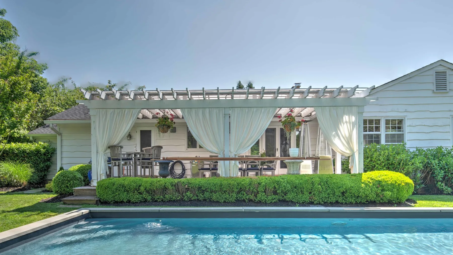 An outdoor pool sits beside a covered patio with seating and white curtains.