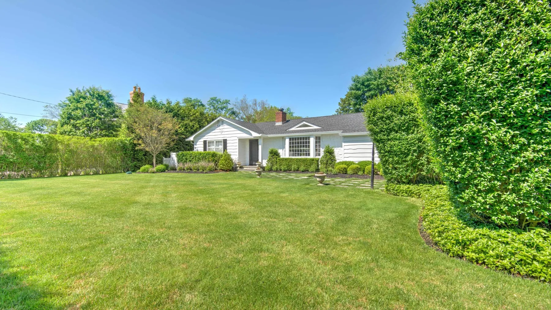 A single-story house sits behind a broad lawn, with trees and hedges around it.