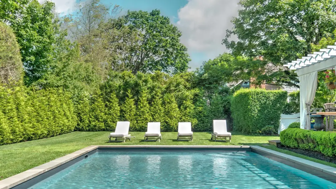 A backyard swimming pool is surrounded by green hedges and lounge seating.