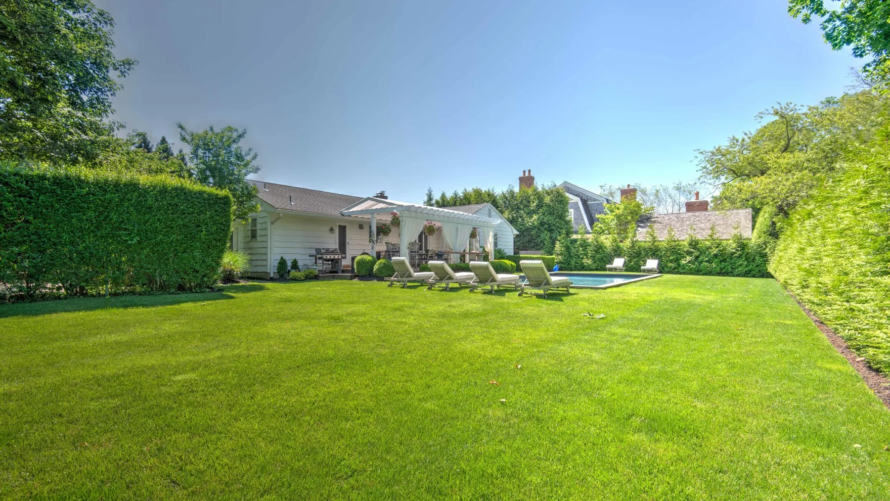 A sunny backyard lawn with hedges, outdoor seating, and a swimming pool.
