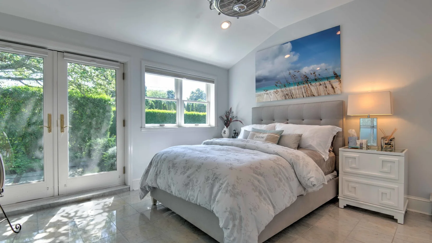 A bright bedroom with a neatly made bed, large windows, and a bedside table.