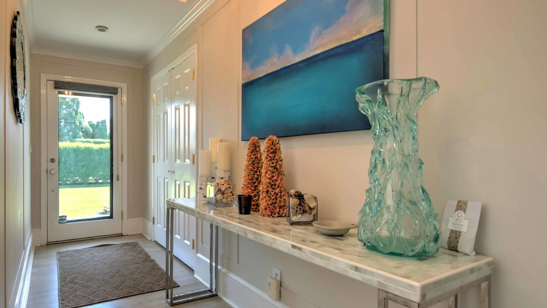 A bright entryway with a glass door and a long console beneath a seascape painting.