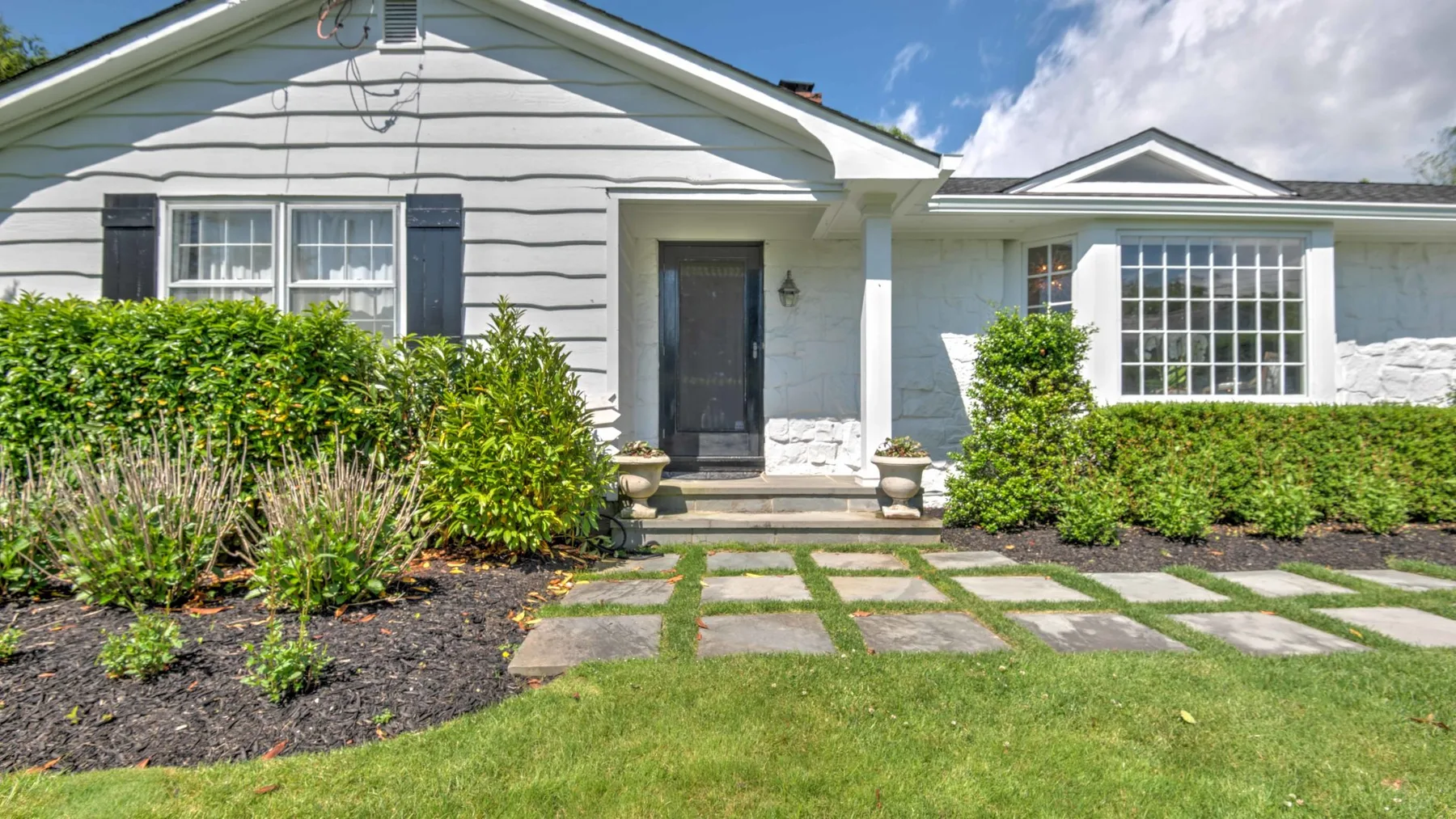 A white house entrance with a dark front door, windows, and garden path.