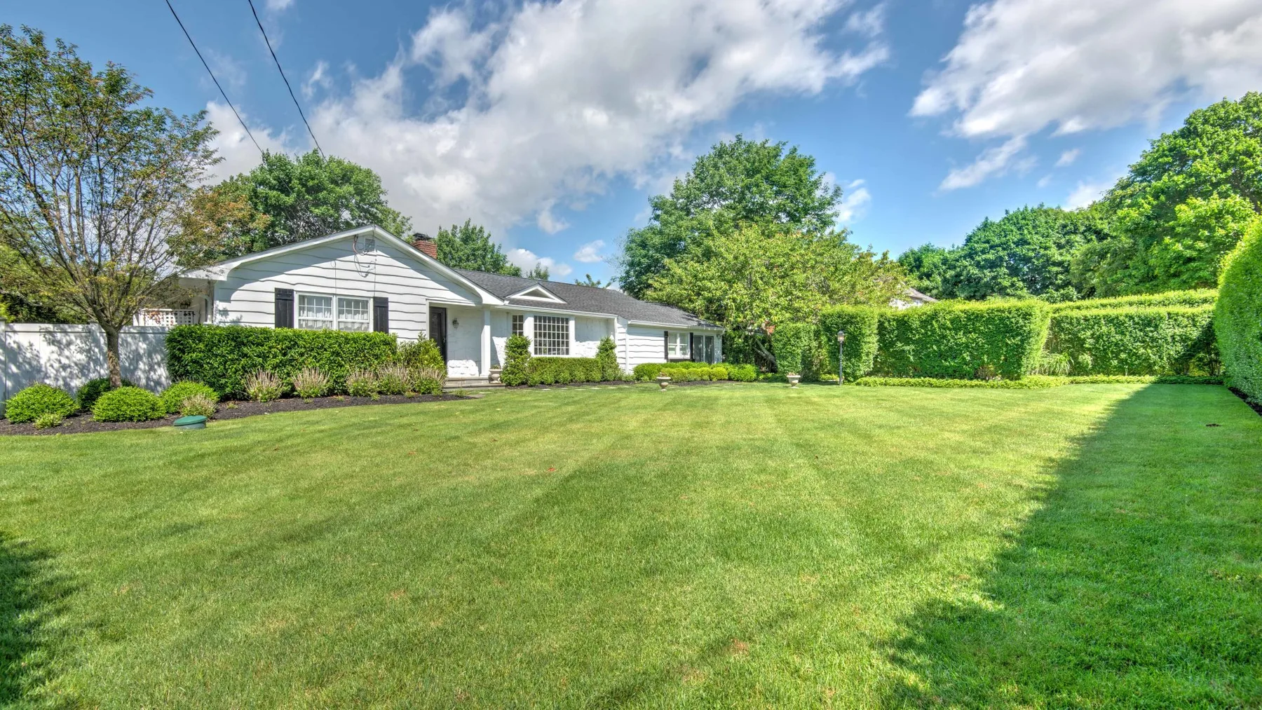 A single-story house sits behind a broad green lawn and leafy hedges.