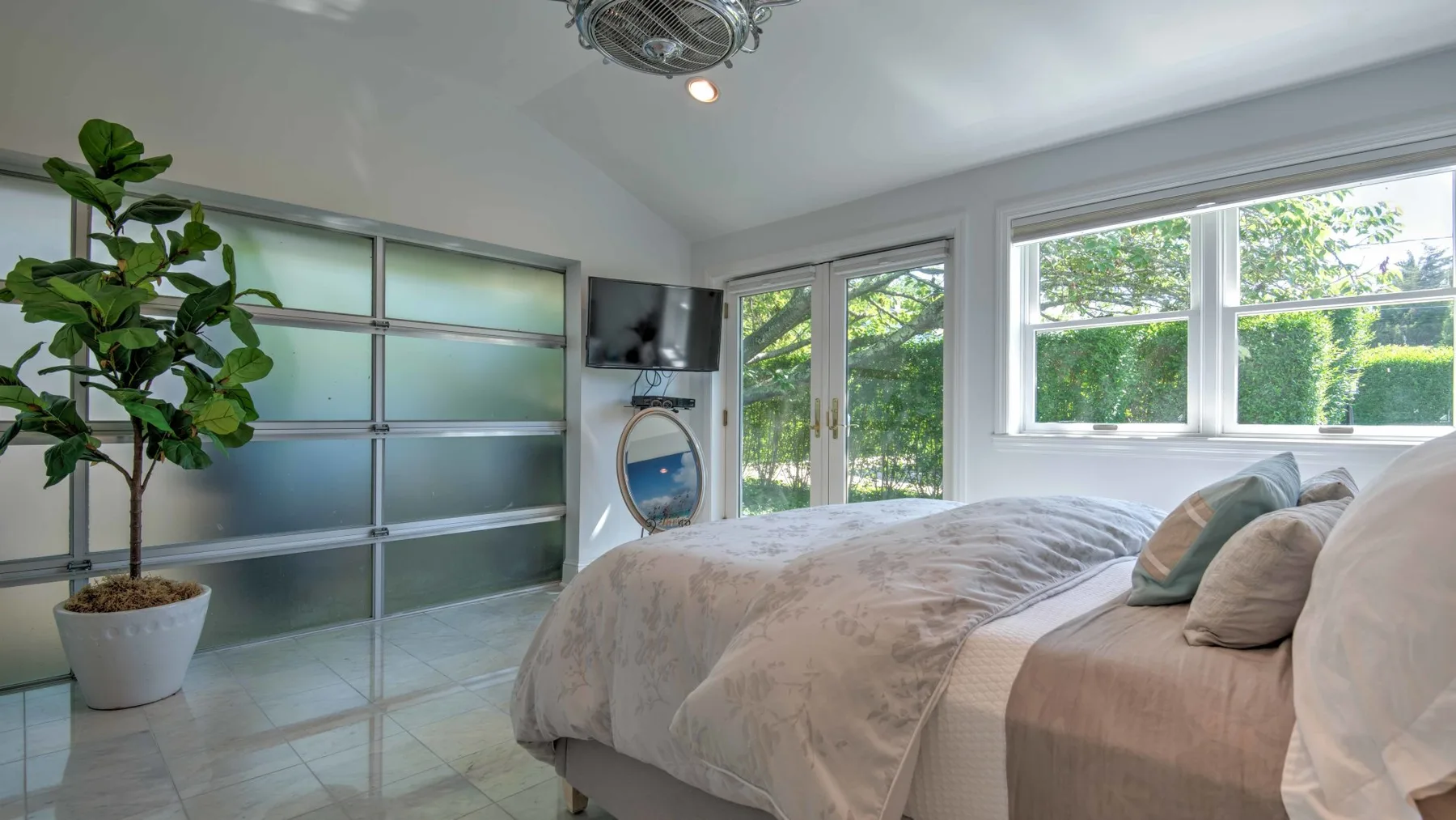 A bright bedroom with a bed beside large windows overlooking greenery.