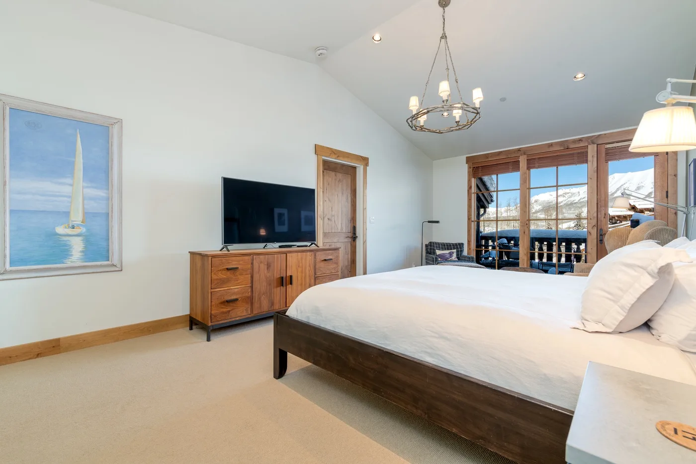 A bright bedroom features a large bed, wooden dresser, and windows overlooking the water.
