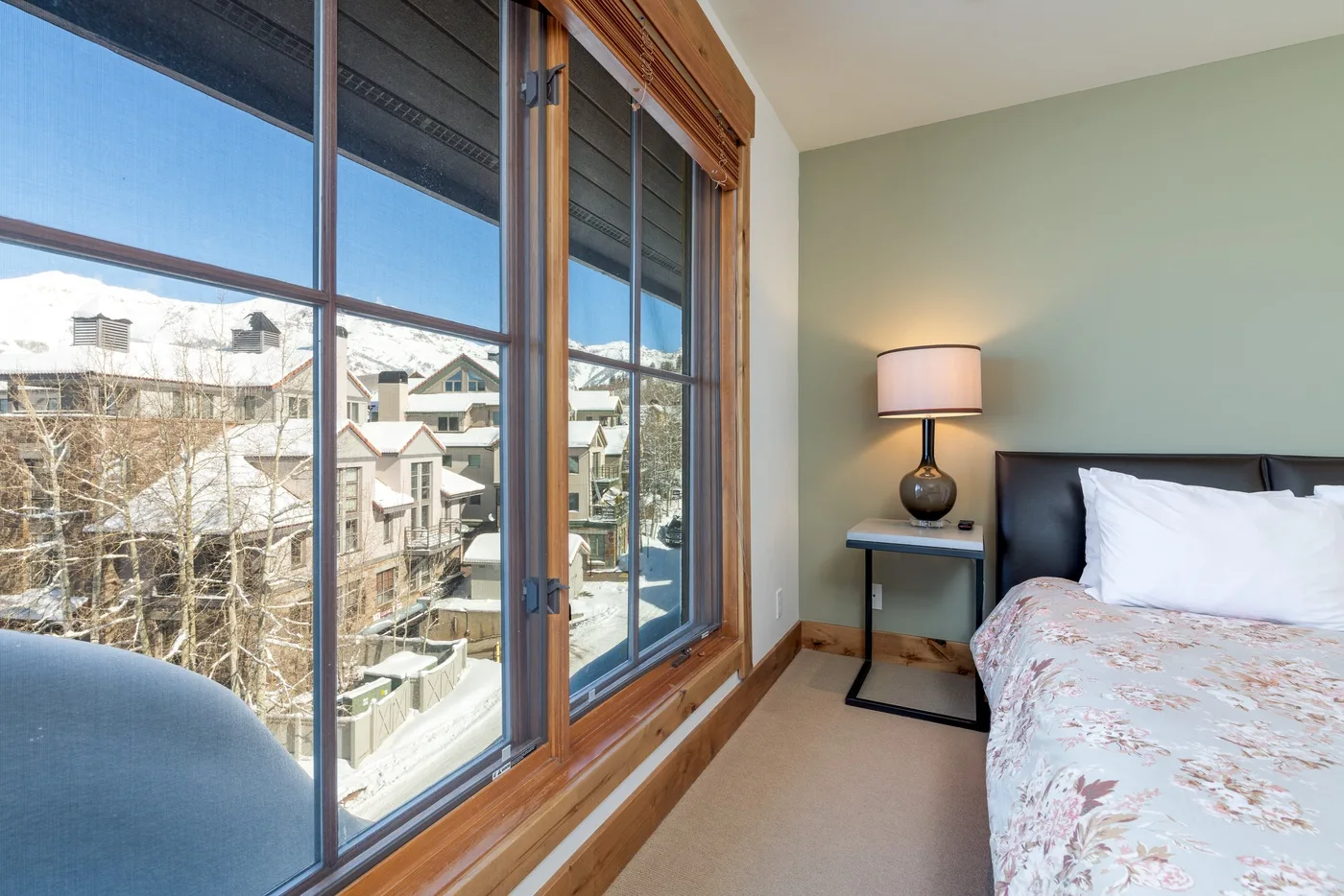 A bedroom with a bed beside large windows overlooking neighboring buildings.