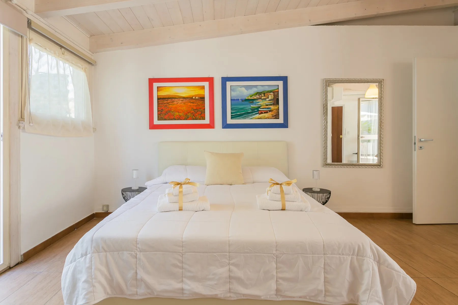 Un dormitorio luminoso con una cama bien tendida y cuadros coloridos sobre ella.