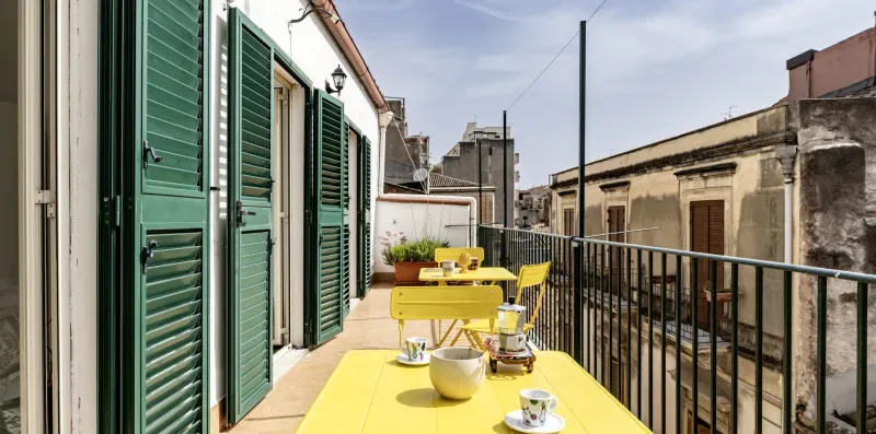 Una terraza en la azotea con una mesa amarilla y sillas junto a contraventanas verdes.