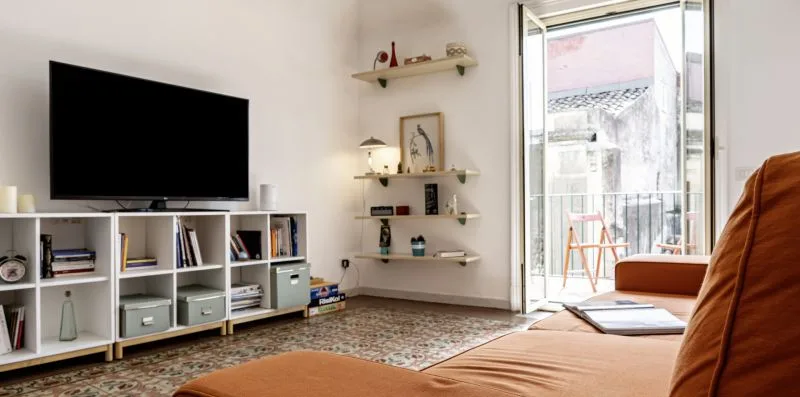 Una sala de estar con televisión, estanterías y una puerta abierta al balcón.