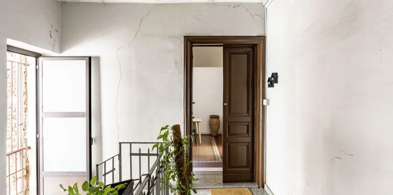 Un pasillo interior luminoso con una puerta abierta y plantas junto a la barandilla.