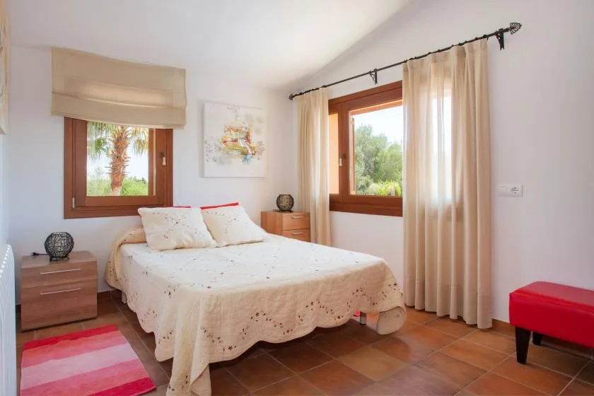 A bedroom with a double bed between two windows and a red bench.