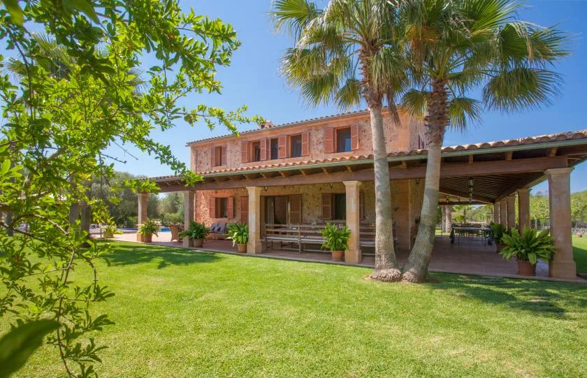 A stone house with a covered patio and palm trees beside a green lawn.