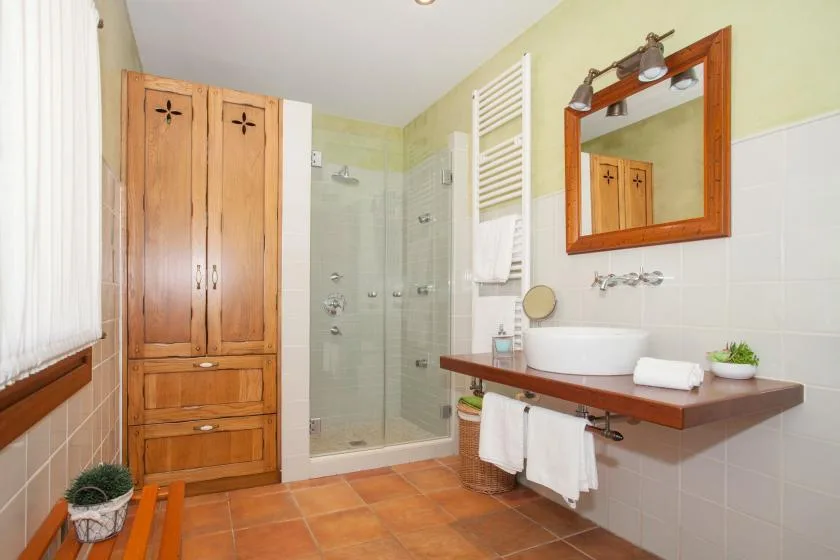 A bathroom with a glass shower, wooden vanity, and terracotta-colored floor tiles.
