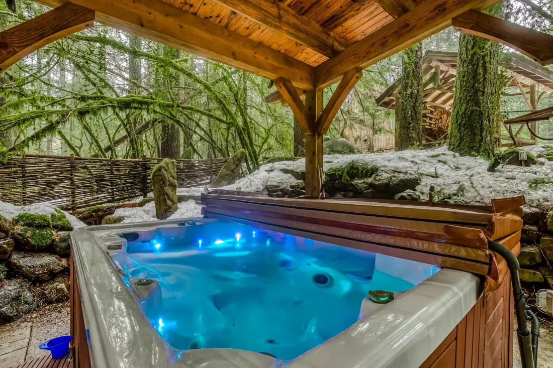 An outdoor hot tub glows beneath a wooden shelter in a snowy woodland setting.
