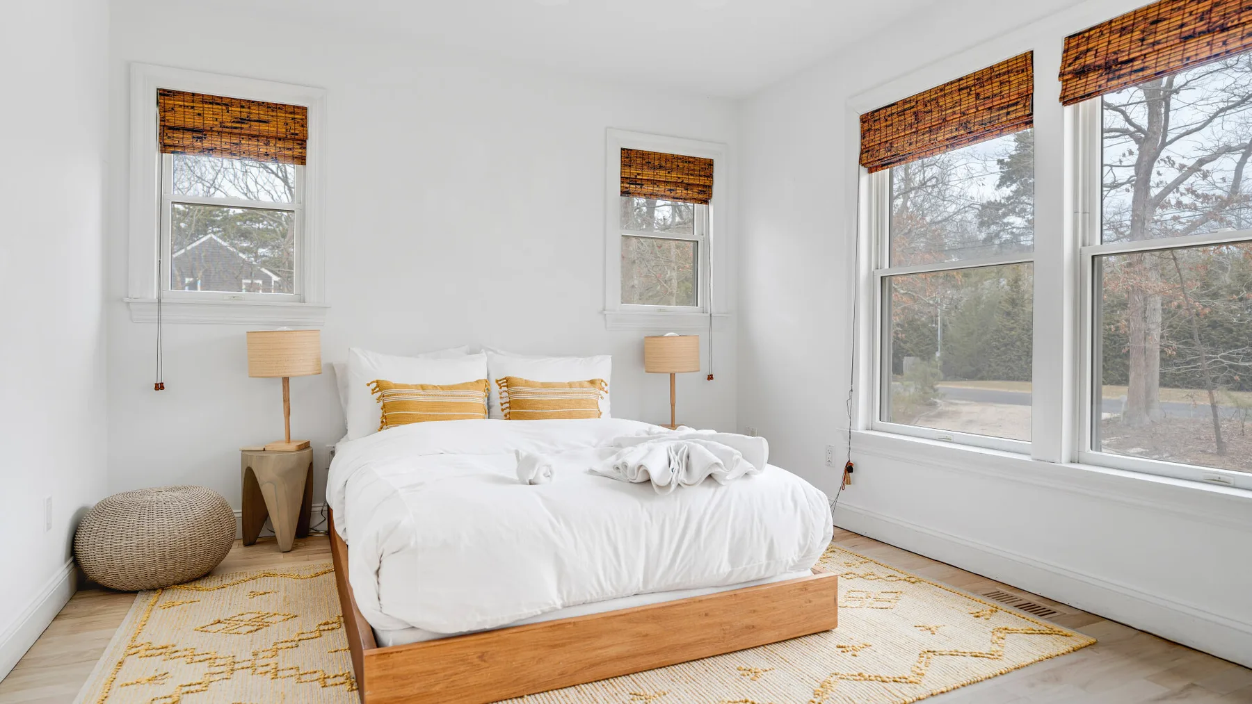 Un dormitorio luminoso con una cama blanca, mesitas de noche y ventanas con vista a los árboles.