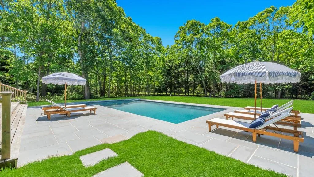 Una piscina rodeada por un patio pavimentado, tumbonas y césped verde.