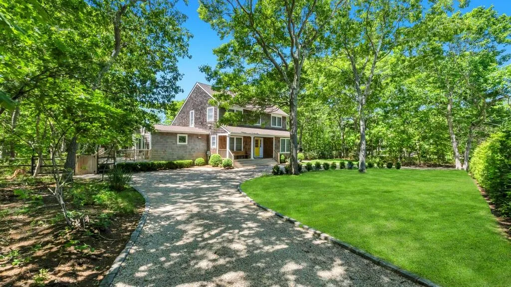 Una casa está junto a una entrada curva y un césped verde, bajo árboles frondosos.