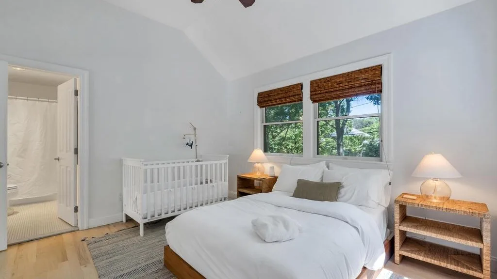 Un dormitorio luminoso con una cama, una cuna y ventanas con vistas a árboles frondosos.