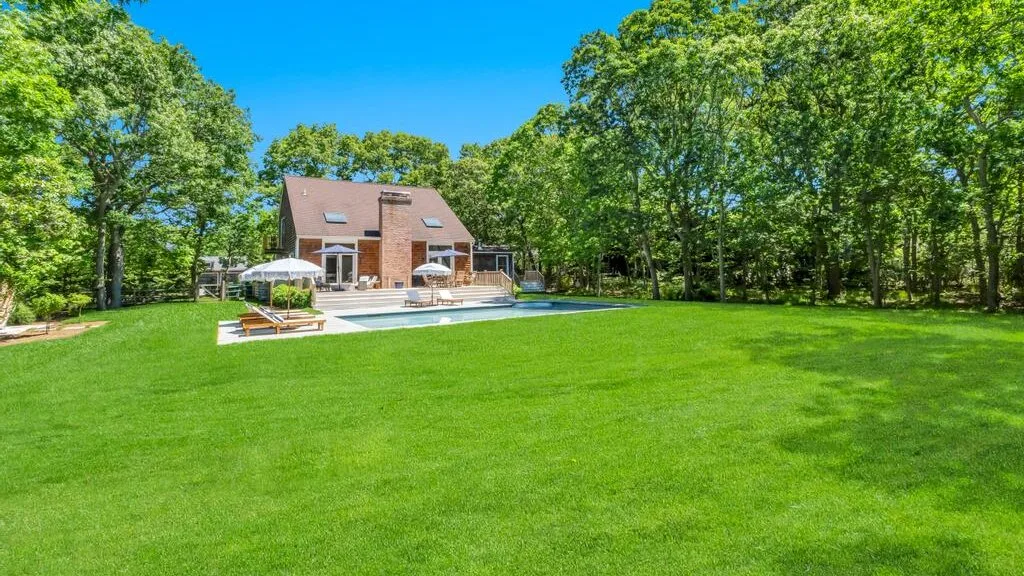 Una casa y asientos al aire libre junto a una piscina en un amplio césped verde.