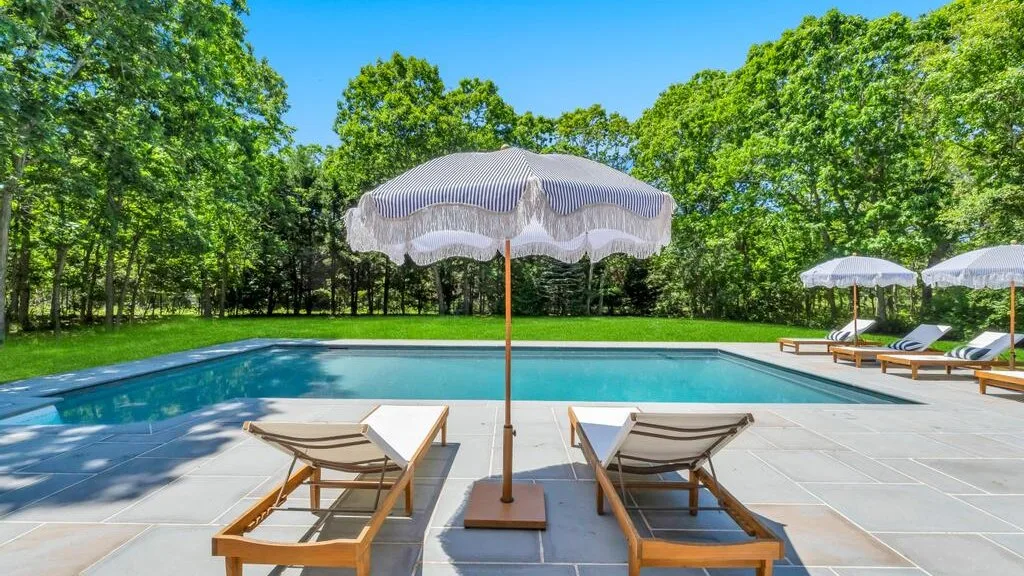 Una piscina al aire libre está rodeada por una terraza pavimentada y árboles frondosos.