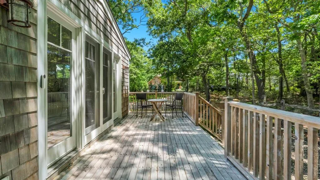Una terraza de madera con asientos al aire libre junto a una casa, rodeada de árboles frondosos.